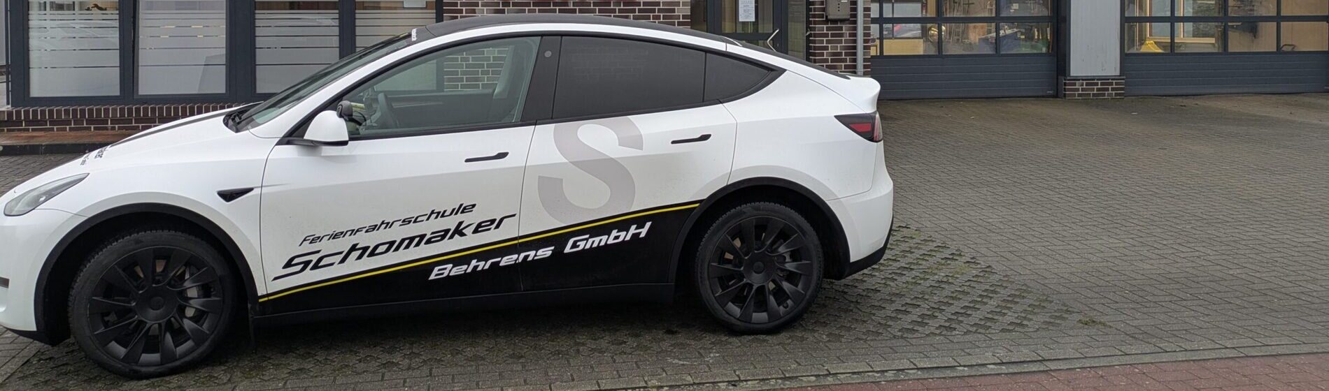 Fahrschulwagen der Fahrschule Schomaker-Behrens — weißer Tesla Model Y mit gelb-schwarzer Beklebung und dem großen S-Signet auf der Tür